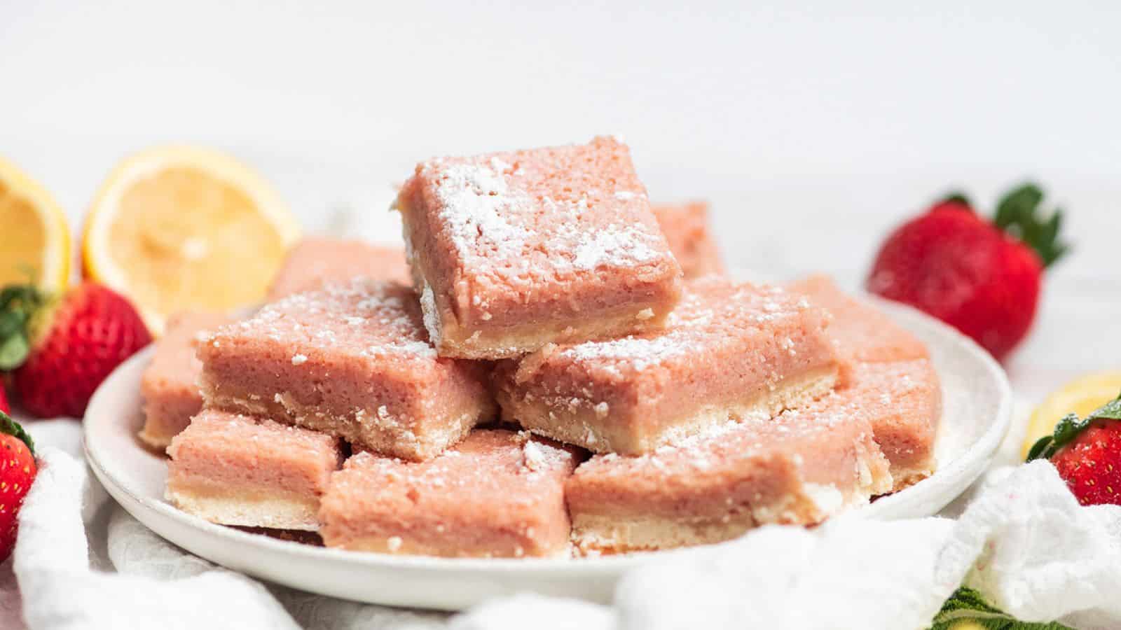A plate of pink strawberry lemon bars dusted with powdered sugar, surrounded by fresh strawberries and lemons.