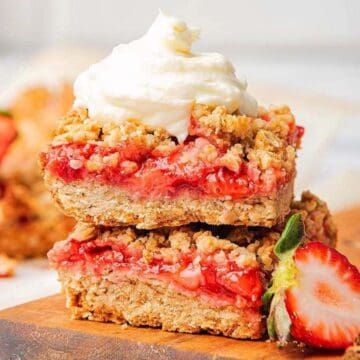 Two strawberry oat bars stacked, topped with whipped cream, with a fresh strawberry on the side.