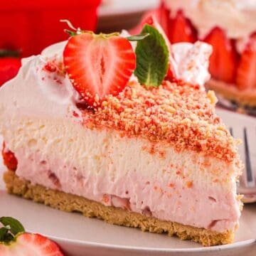 A slice of strawberry shortcake cheesecake topped with whipped cream, crumbs, and fresh strawberries on a plate.