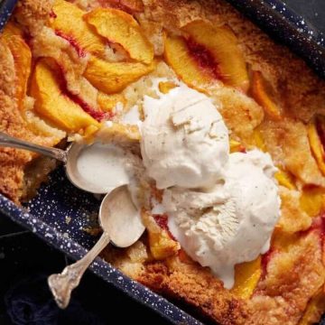 Peach cobbler, inspired by vintage recipes, topped with two scoops of vanilla ice cream and served with two spoons in a dark baking dish.