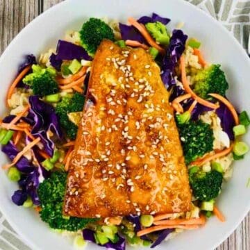 Bowl of glazed salmon on rice with broccoli, shredded carrots, cabbage, and sesame seeds, with a fork on the side.