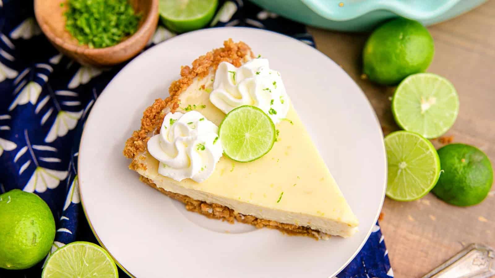 A slice of key lime pie topped with whipped cream and lime, surrounded by whole and sliced limes on a plate.