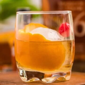 A cocktail with orange peel, ice, and a cherry in a glass on a wooden surface.