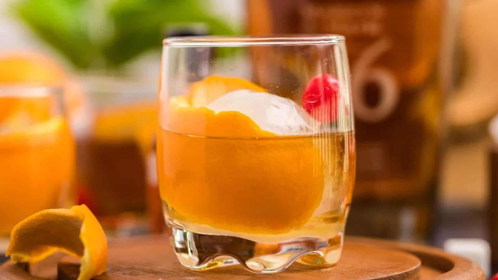 A cocktail with orange peel, ice, and a cherry in a glass on a wooden surface.