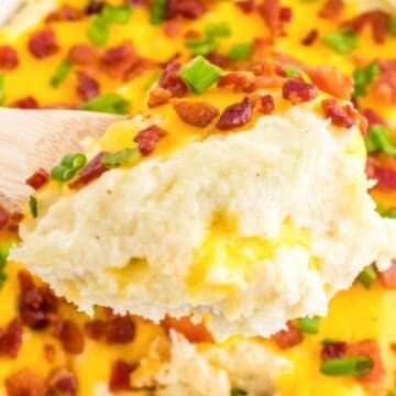 A wooden spoon holds cheesy mashed potatoes topped with bacon bits and chopped green onions.