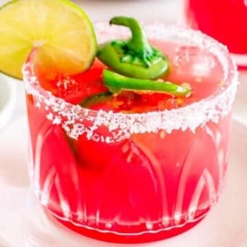 A bright red cocktail in a salt-rimmed glass, garnished with lime and jalapeño slices.