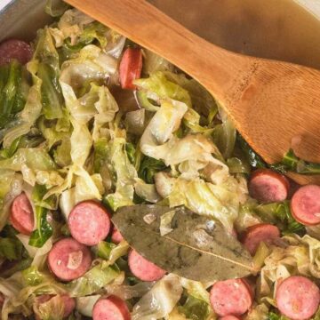Sliced sausage and cabbage cooked in a pot with broth, garnished with a bay leaf and stirred with a wooden spoon.
