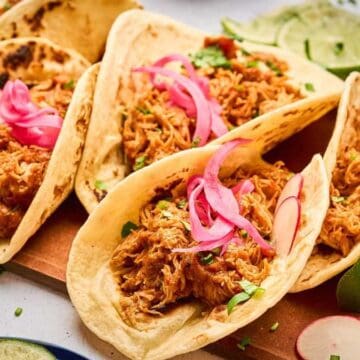 Four tacos filled with shredded meat, pickled onions, and radishes on a wooden board with lime wedges.