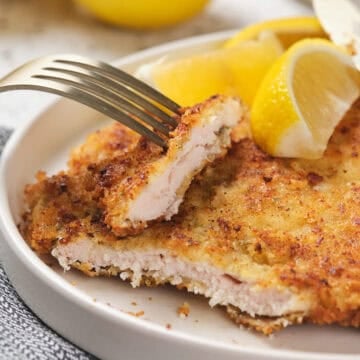 A fork and knife cut into a breaded chicken cutlet with lemon wedges on a white plate.