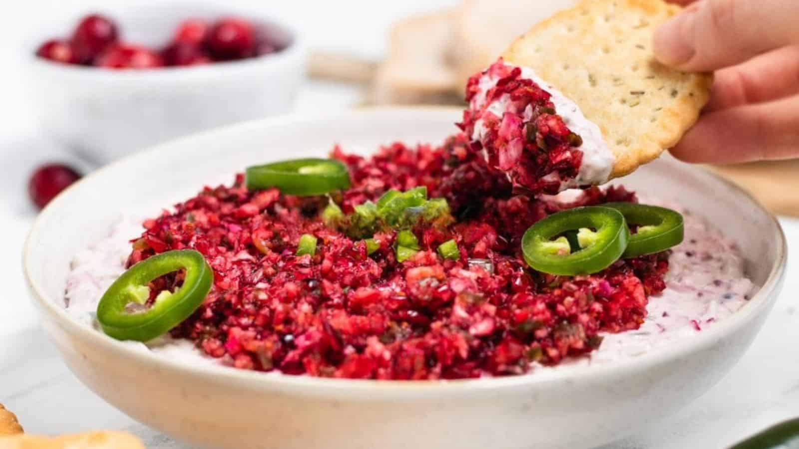 A cracker is dipped into a bowl of creamy cranberry jalapeño dip, garnished with sliced jalapeños.