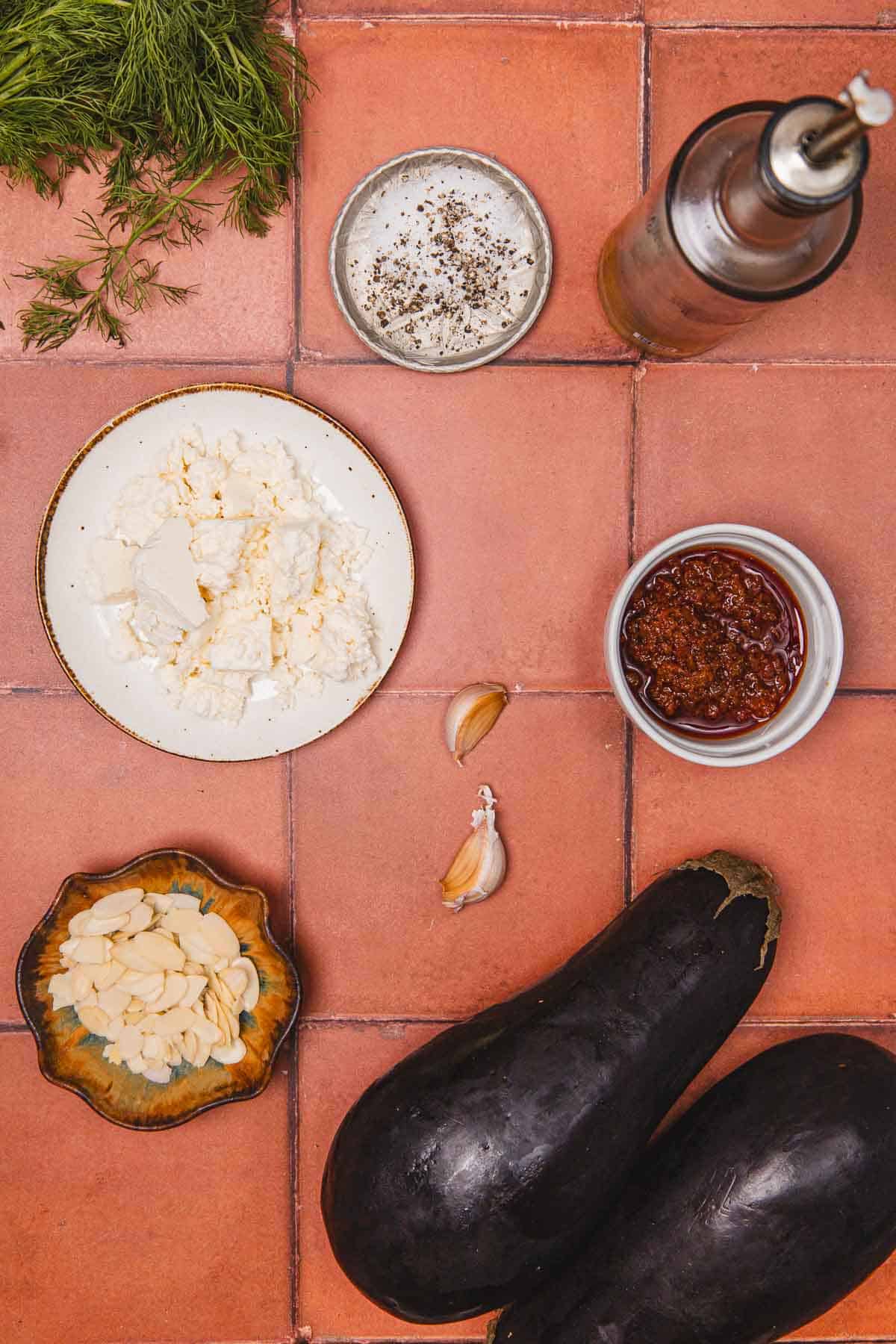 Ingredients on a tiled surface: eggplants, feta, garlic, oil, dill, chili paste, sliced almonds, salt, and pepper.