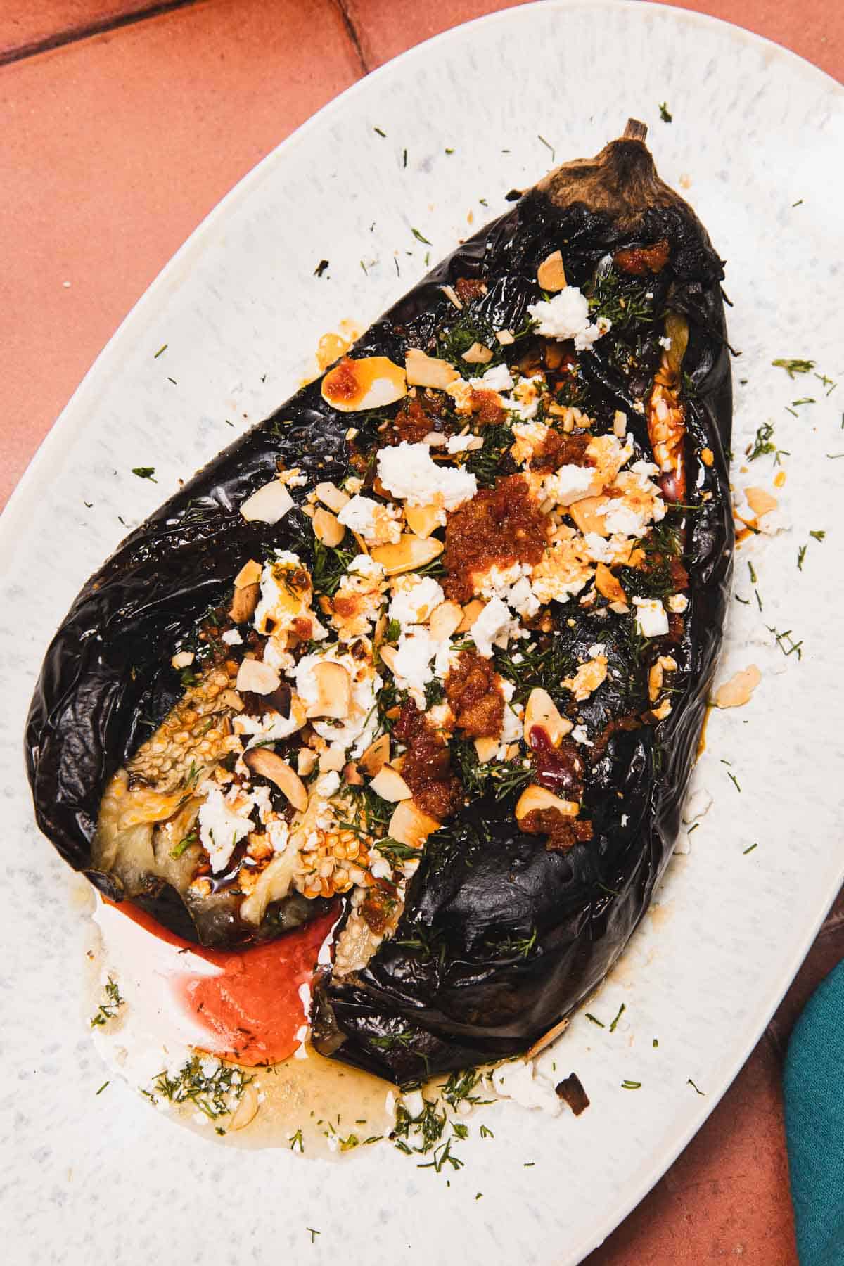A roasted eggplant topped with crumbled cheese, nuts, and herbs on a white oval plate.