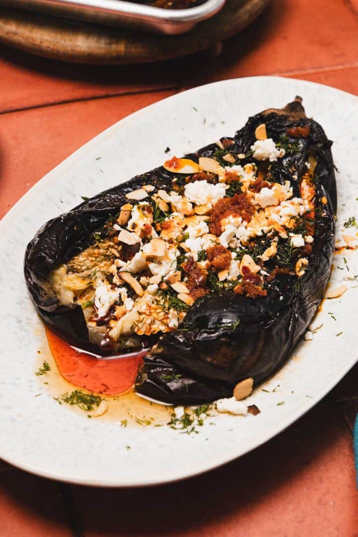 Roasted eggplant topped with crumbled cheese, herbs, nuts, and sauce on a white oval plate.