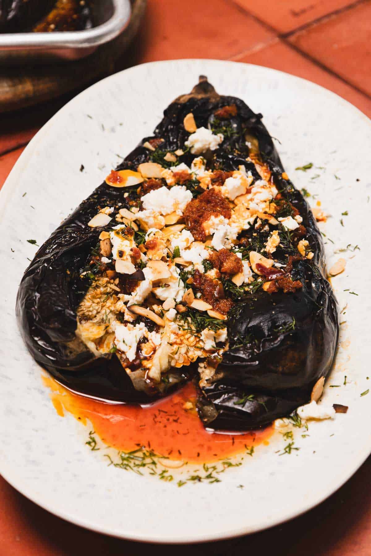 A roasted eggplant topped with crumbled cheese, nuts, herbs, and red sauce on a white plate.