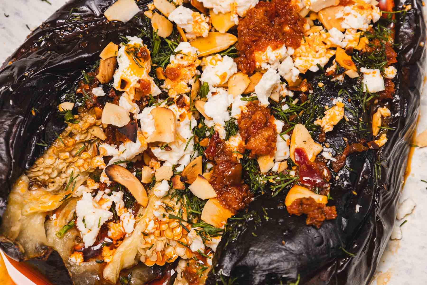 Close-up of a roasted eggplant topped with crumbled cheese, herbs, tomato sauce, and sliced almonds.