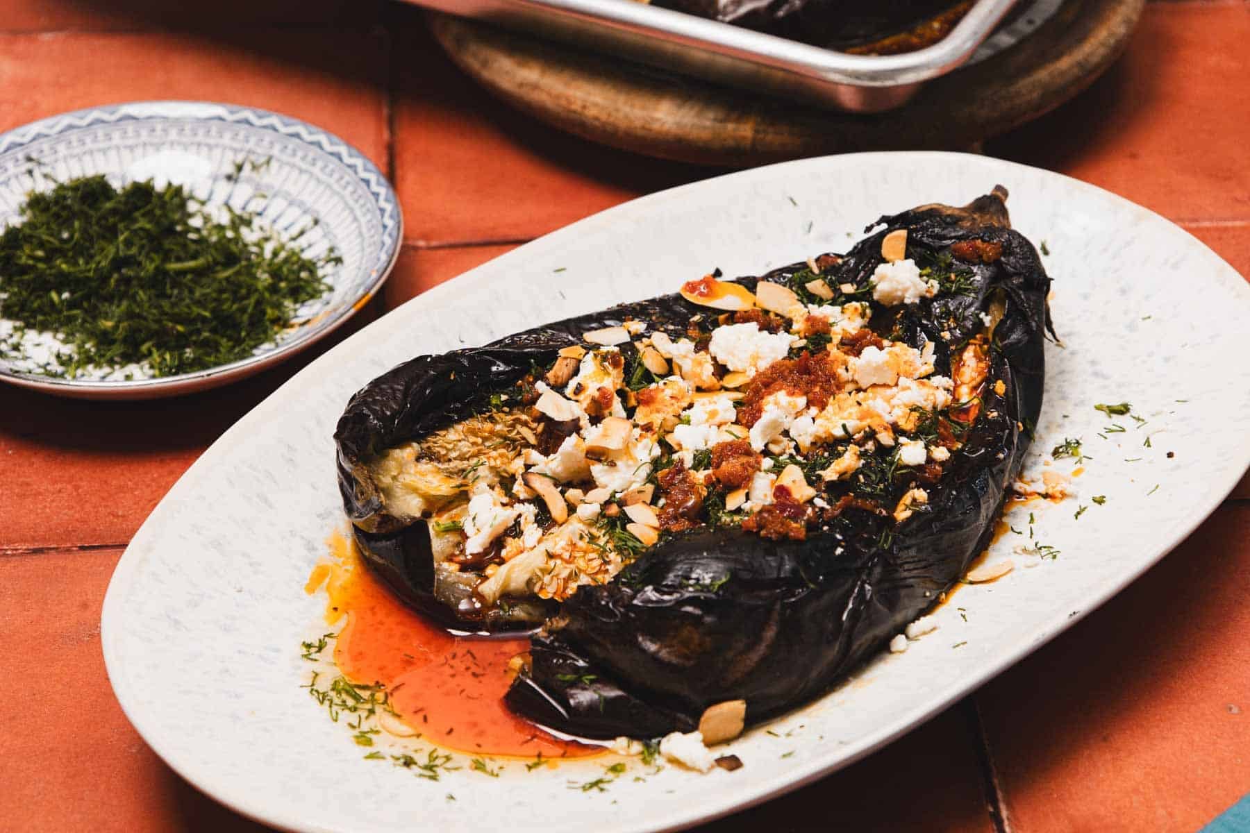 Roasted eggplant topped with crumbled cheese, herbs, and spices on a white plate with a side of chopped herbs.
