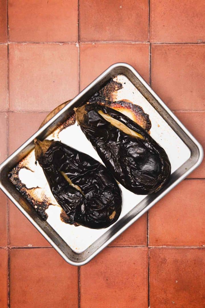 Two roasted eggplants on a baking tray with burnt marks, placed on a terracotta tiled surface.