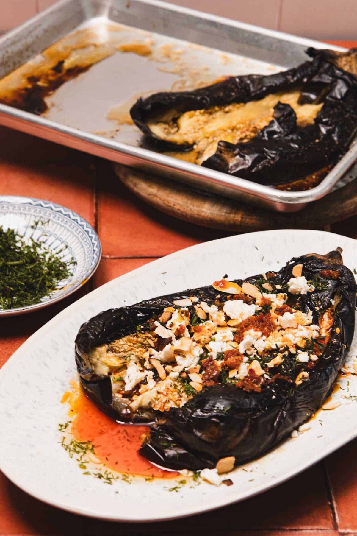 Roasted eggplant topped with crumbled cheese, herbs, and spices on a plate, with another on a baking tray.