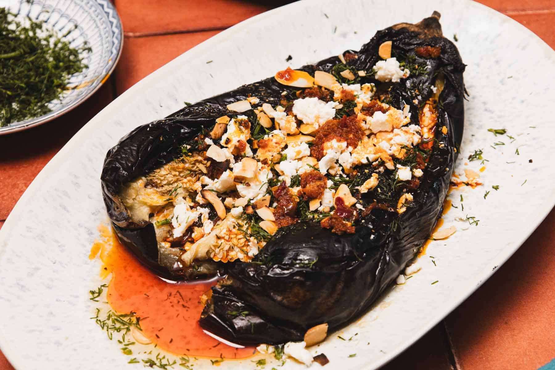 Roasted eggplant topped with feta cheese, nuts, herbs, and red sauce on an oval white plate.