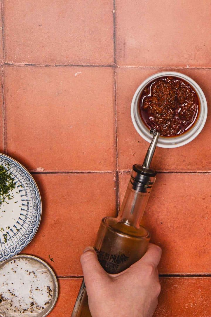 A hand pours olive oil into a small bowl of chili sauce on a terracotta tile surface.