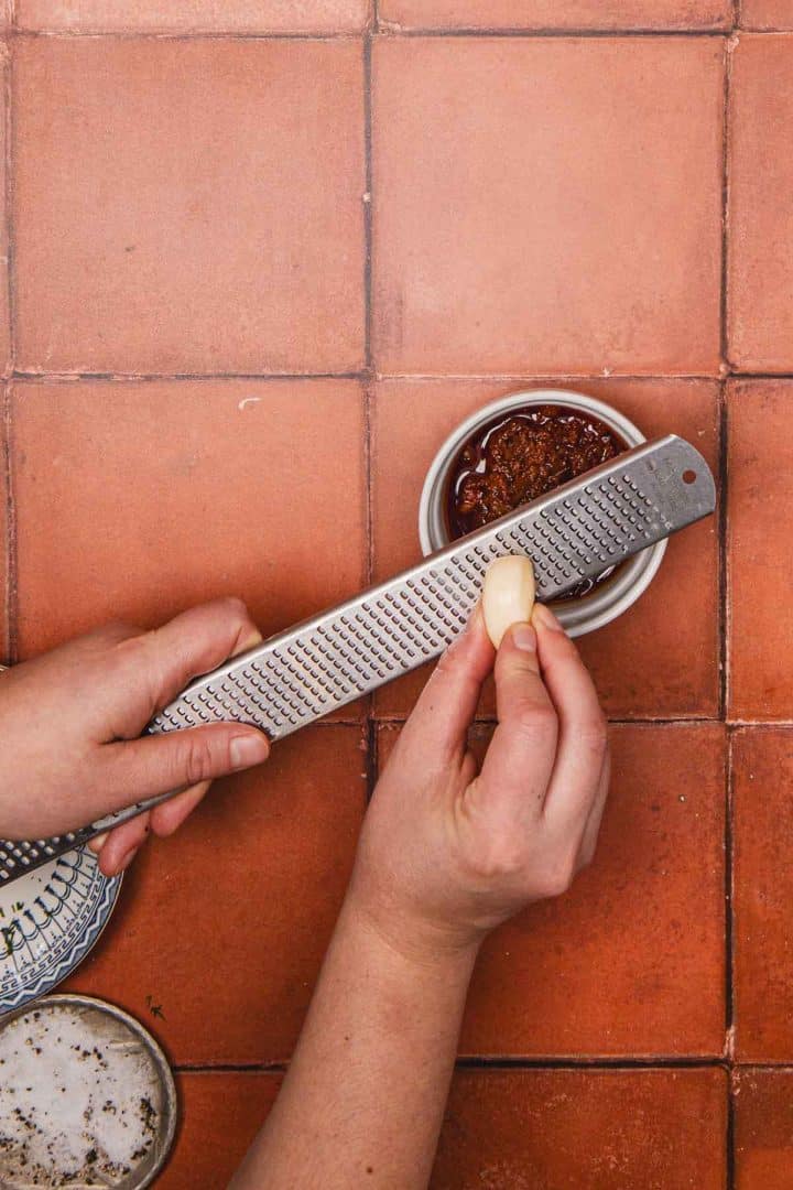 A hand grates garlic over a bowl of sauce on a terracotta-tiled surface.