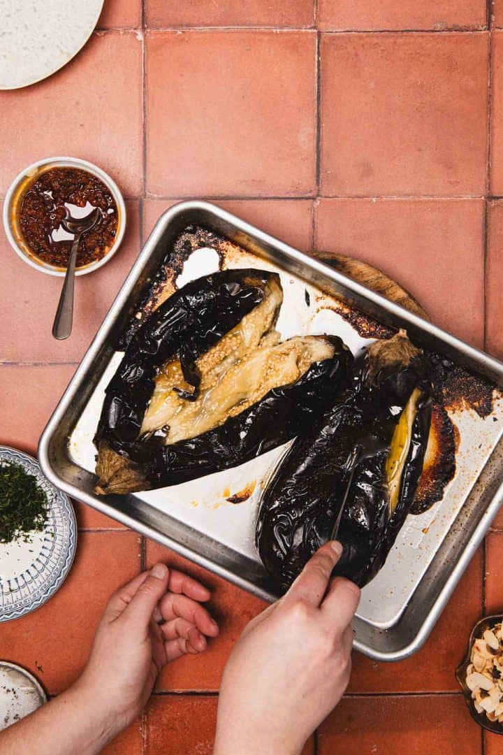 Two roasted eggplants on a baking tray, with a hand scooping the flesh, surrounded by small bowls on a tile surface.
