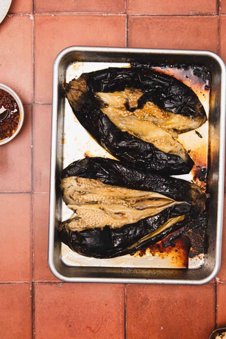 Charred roasted eggplants on a metal baking tray, placed on a red tiled surface.