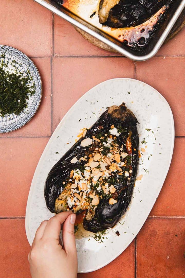 Roasted eggplant topped with feta, almonds, and herbs on a plate; a hand reaches in to take some.