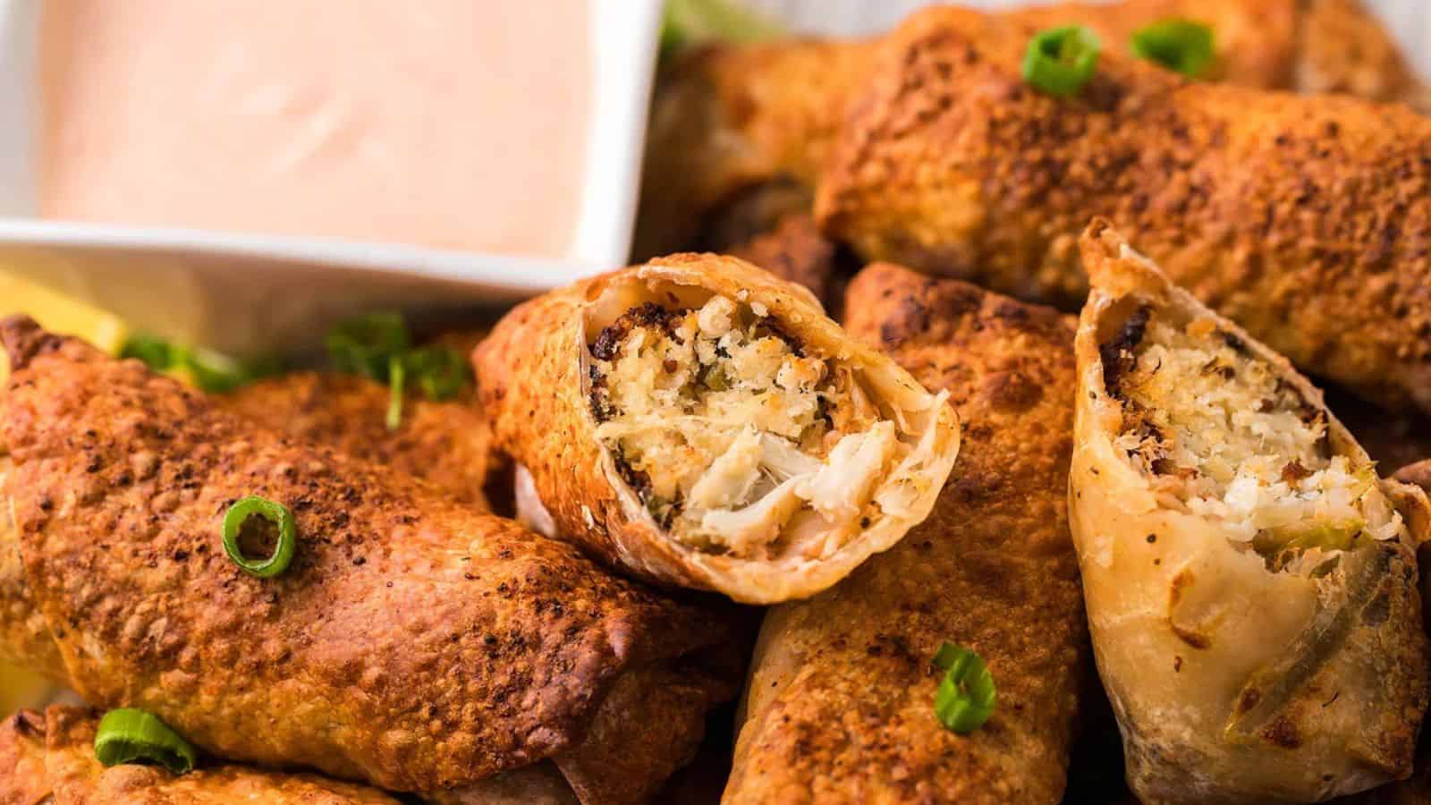 Close-up of crispy egg rolls, one split open to reveal filling, with a creamy dipping sauce in the background.