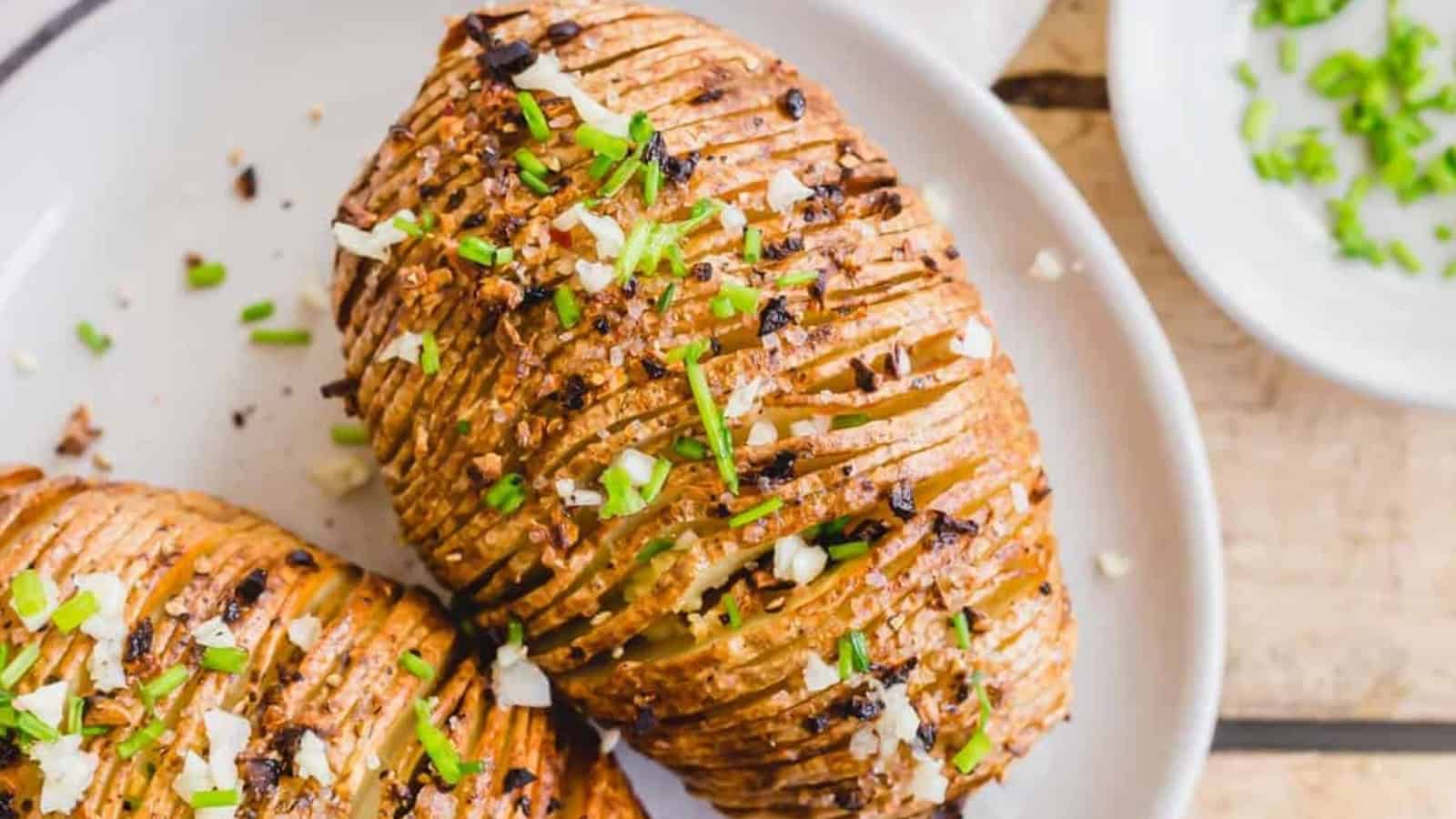 Crispy Hasselback potatoes topped with chopped herbs, garlic, and seasoning on a white plate.