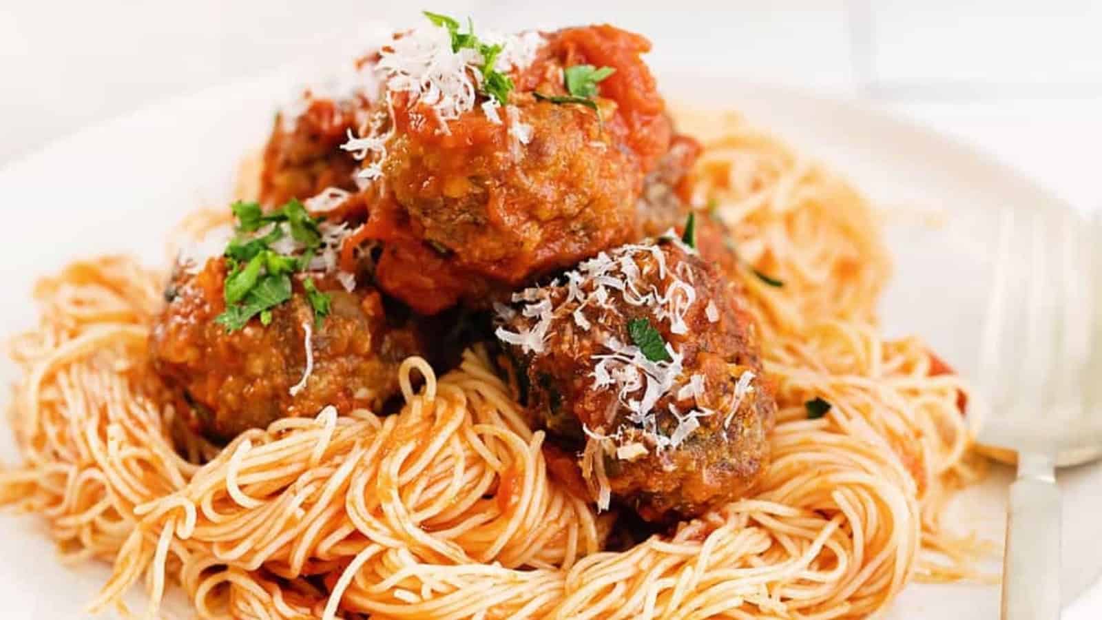 Spaghetti topped with three meatballs, tomato sauce, grated cheese, and chopped parsley on a white plate.