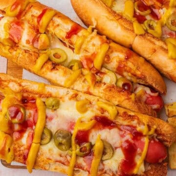 Three hot dogs topped with melted cheese, sliced olives, ketchup, and mustard, served with fries on the side.