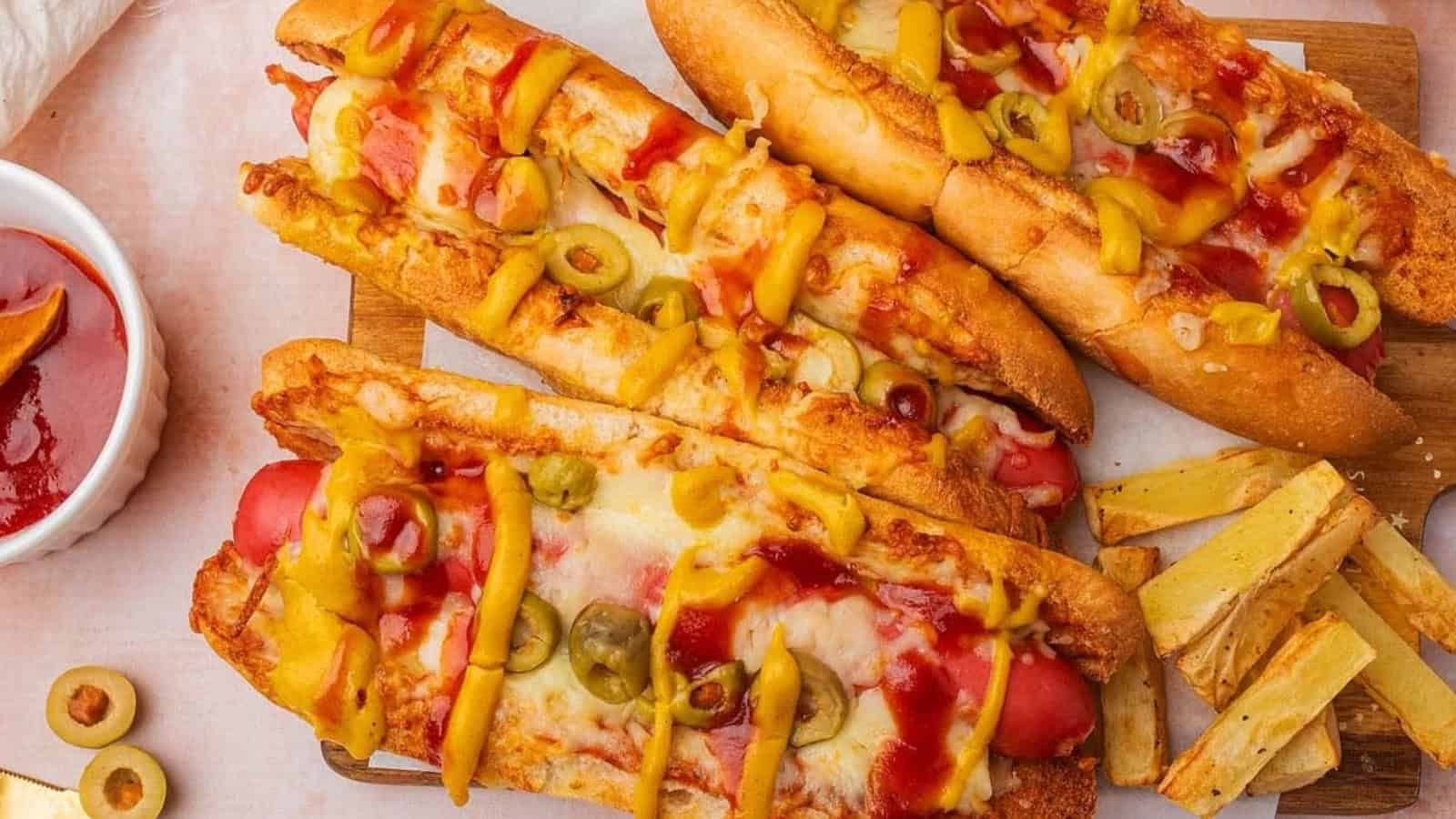 Three hot dogs topped with melted cheese, sliced olives, ketchup, and mustard, served with fries on the side.