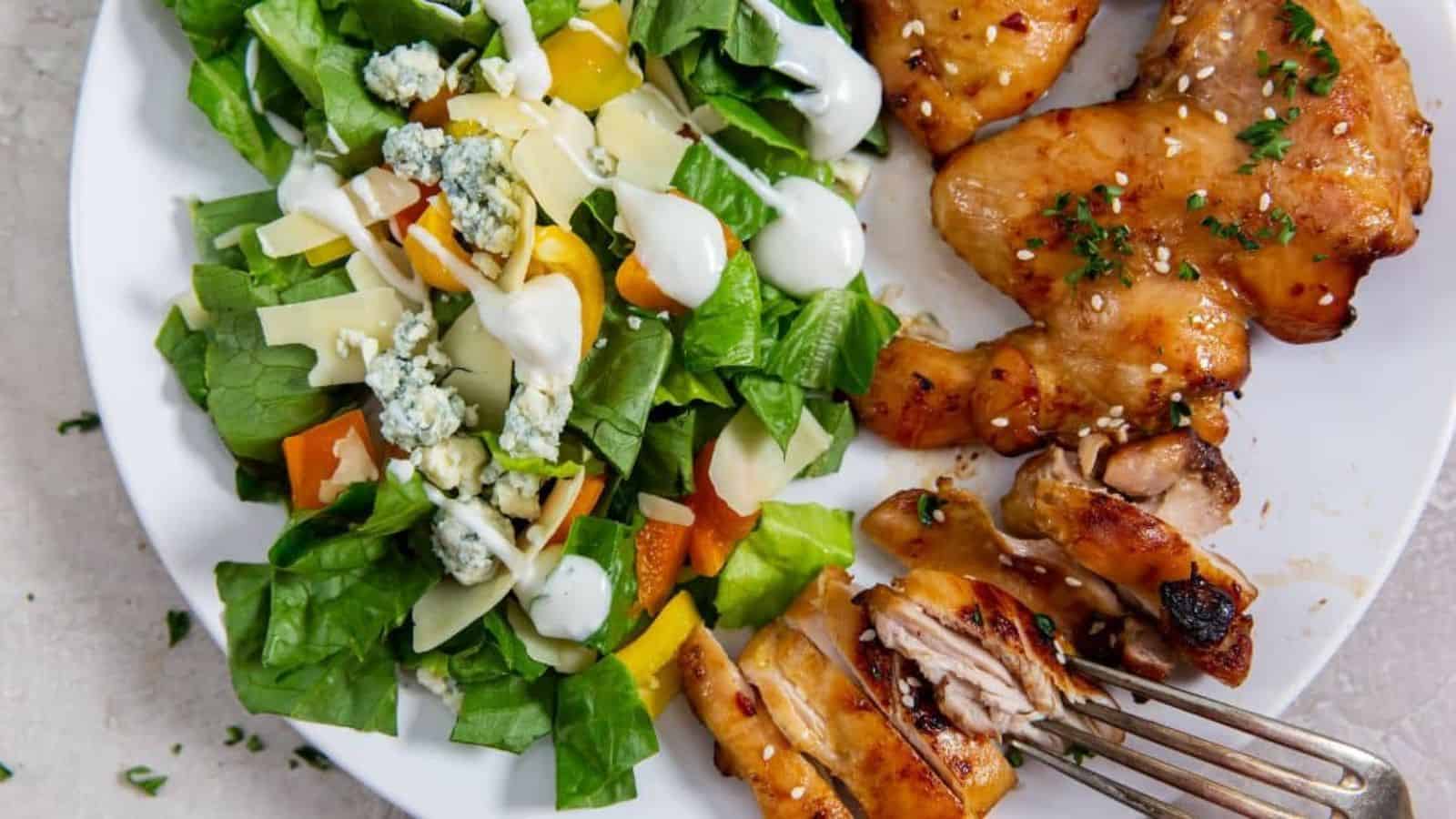 Air Fryer Teriyaki Chicken Thighs Paleo on a white plate with parsley, sesame seeds, lettuce with bell peppers, bleu cheese, parmesan and a fork.