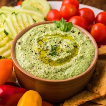 A bowl of green dip with olive oil, surrounded by crackers, cherry tomatoes, cucumber, and mini peppers.