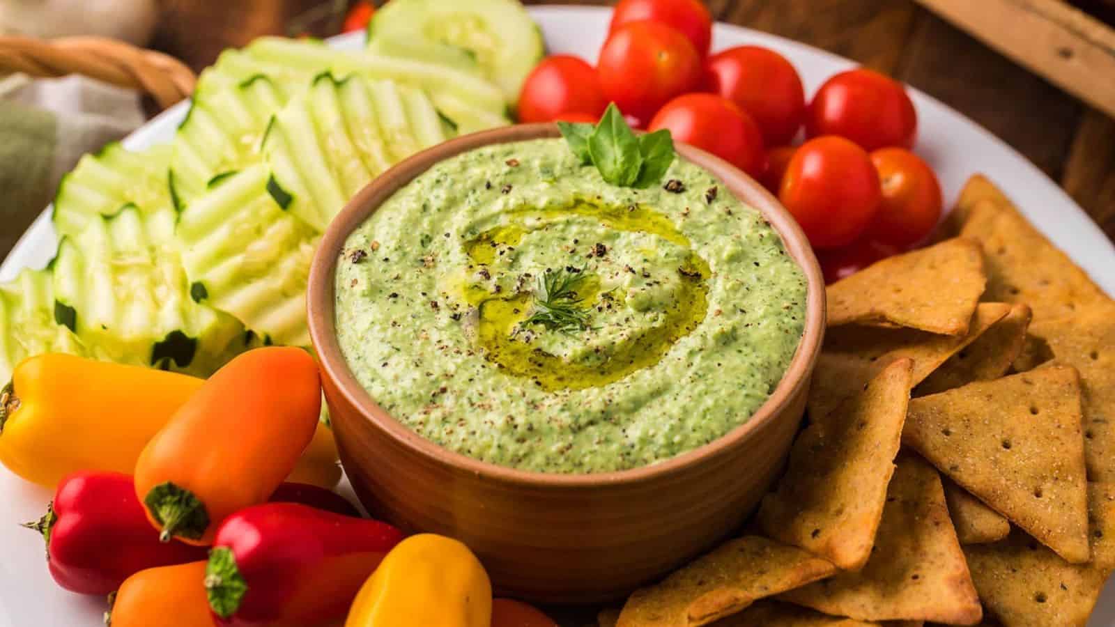 A bowl of green dip with olive oil, surrounded by crackers, cherry tomatoes, cucumber, and mini peppers.