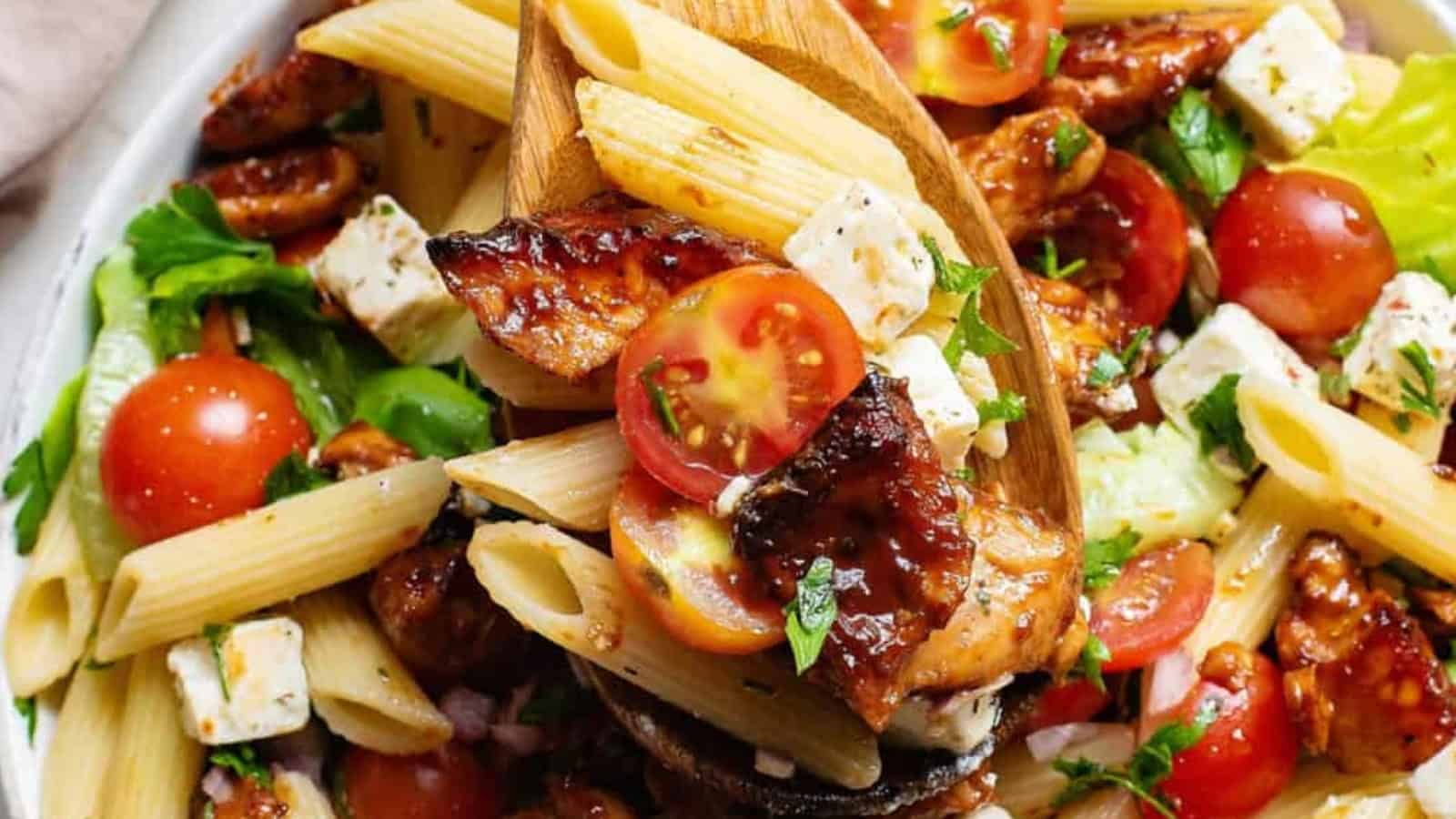 Pasta salad with grilled chicken, cherry tomatoes, feta cheese, and lettuce, served with a wooden spoon.