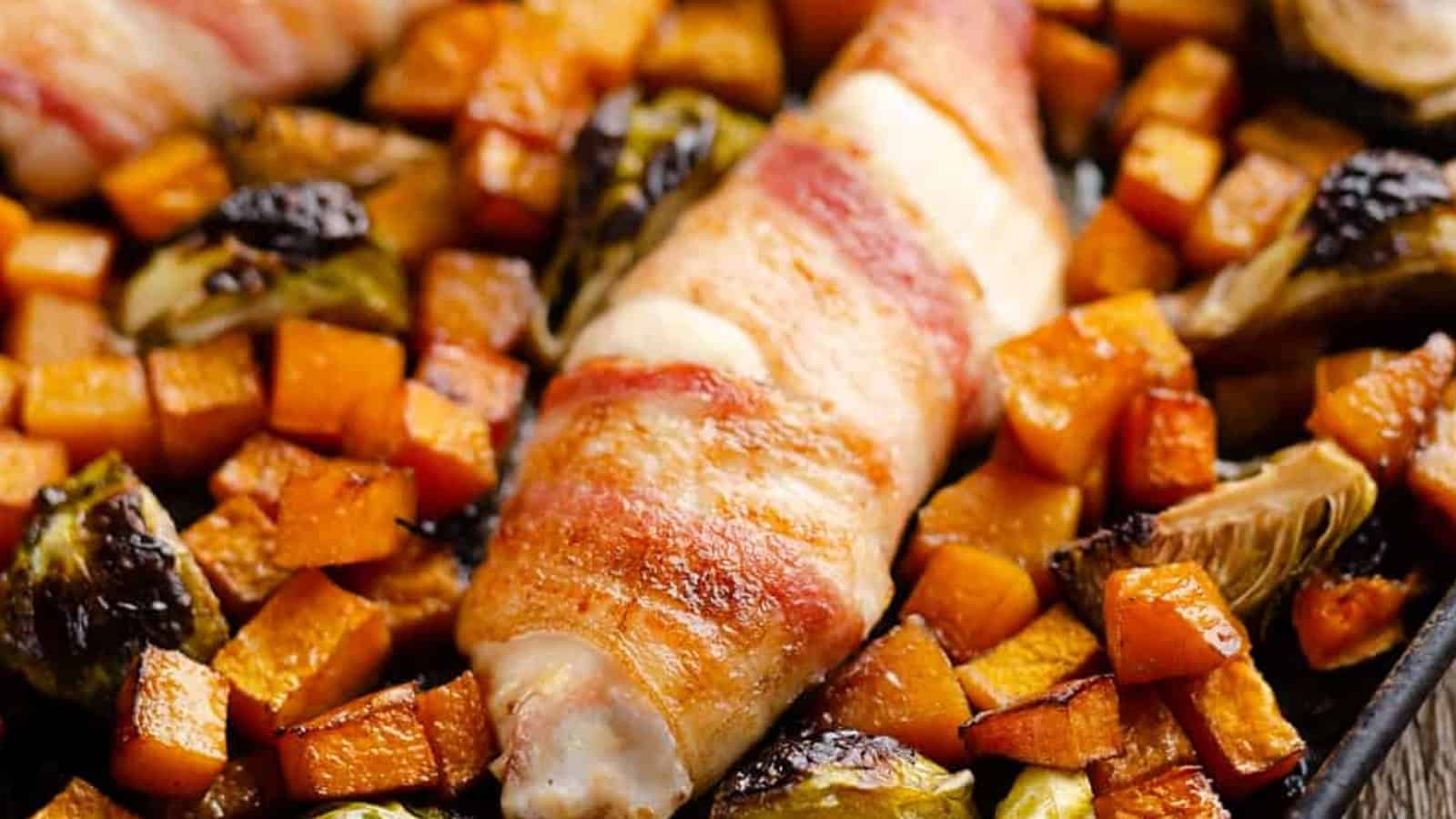 Bacon-wrapped chicken surrounded by roasted butternut squash and brussels sprouts on a baking sheet.