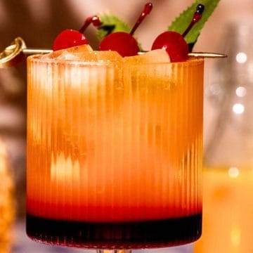 A vibrant cocktail with a sunset gradient, topped with cherries and mint, sits in a ribbed glass.