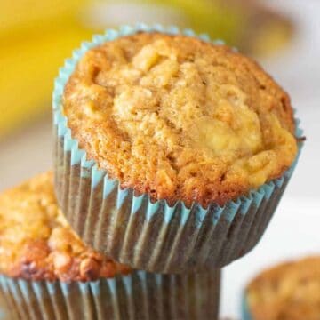 Close-up of banana muffins stacked on a plate with a blurred banana in the background.