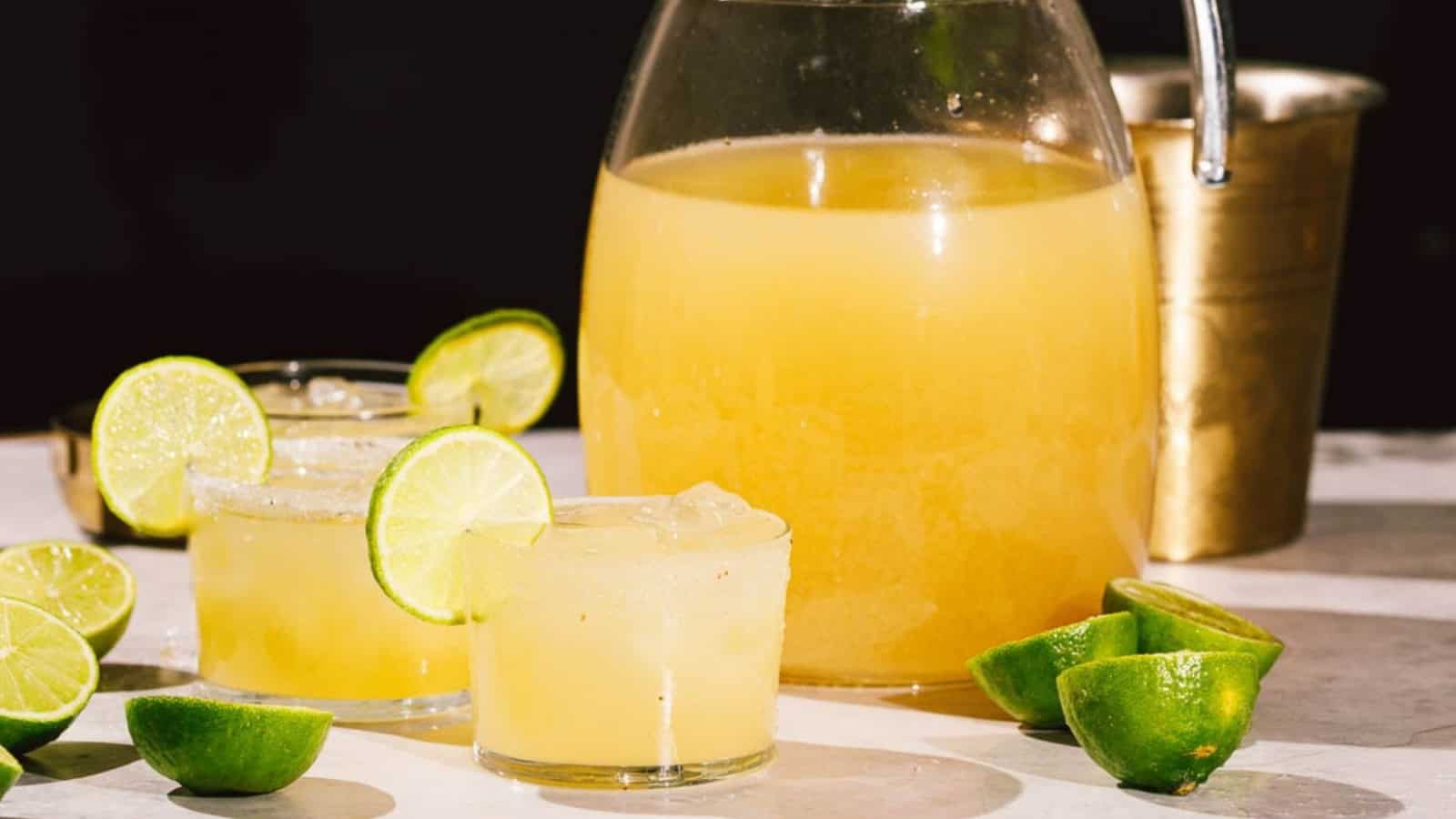 A pitcher and glasses of lime margarita with ice and lime slices, next to cut limes on a white surface.