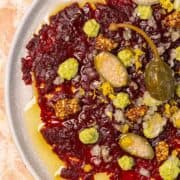 A close-up of a beet carpaccio garnished with capers, mustard seeds, diced onions, and green sauce on a plate.