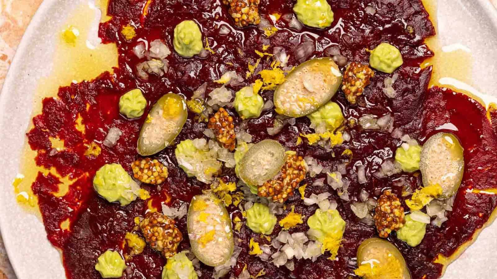 Colorful beet carpaccio topped with green cream, mustard seeds, capers, diced onions, and lemon zest on a plate.