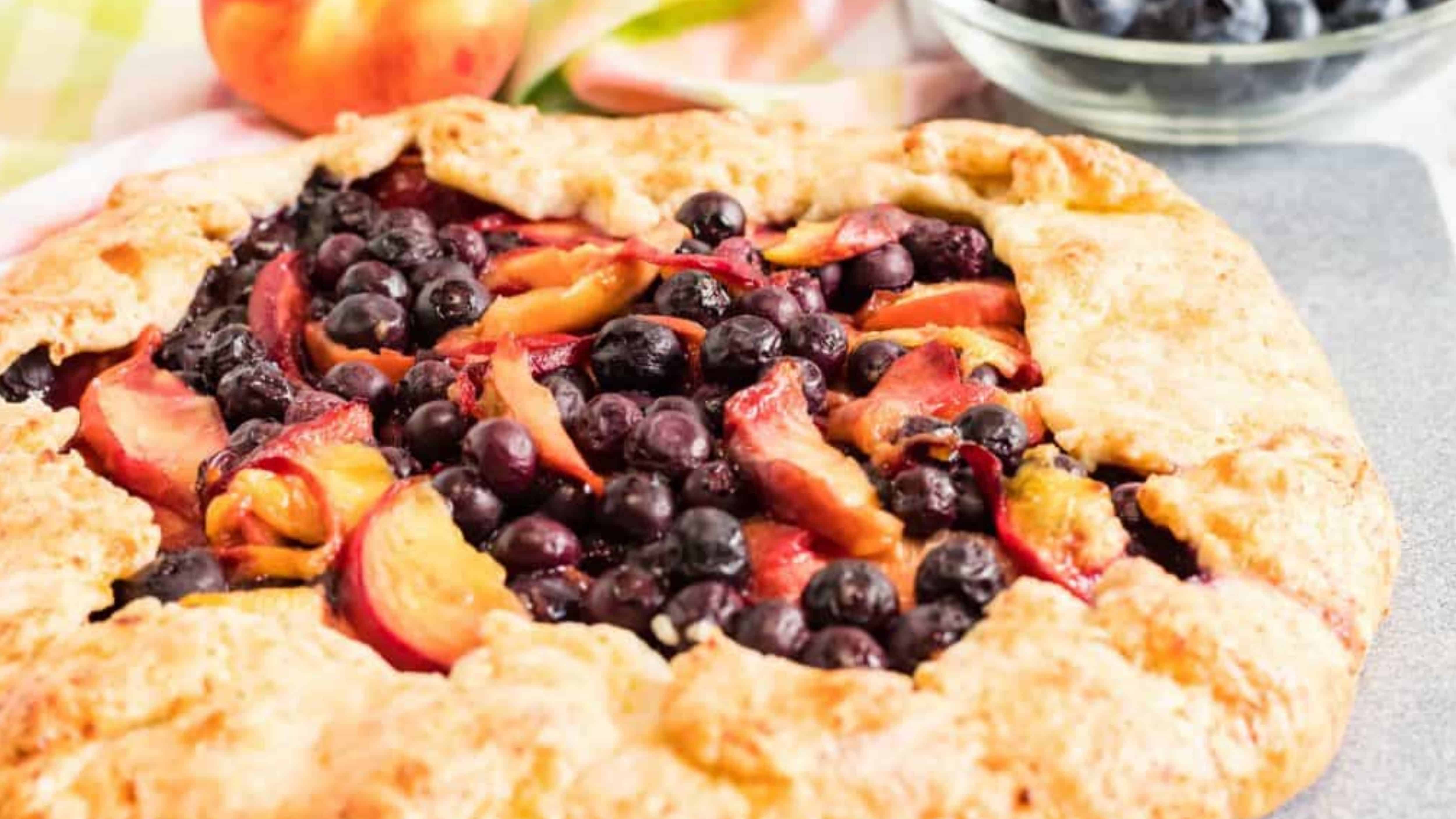 Rustic fruit galette filled with blueberries and peach slices on a table, with more fruit in the background.