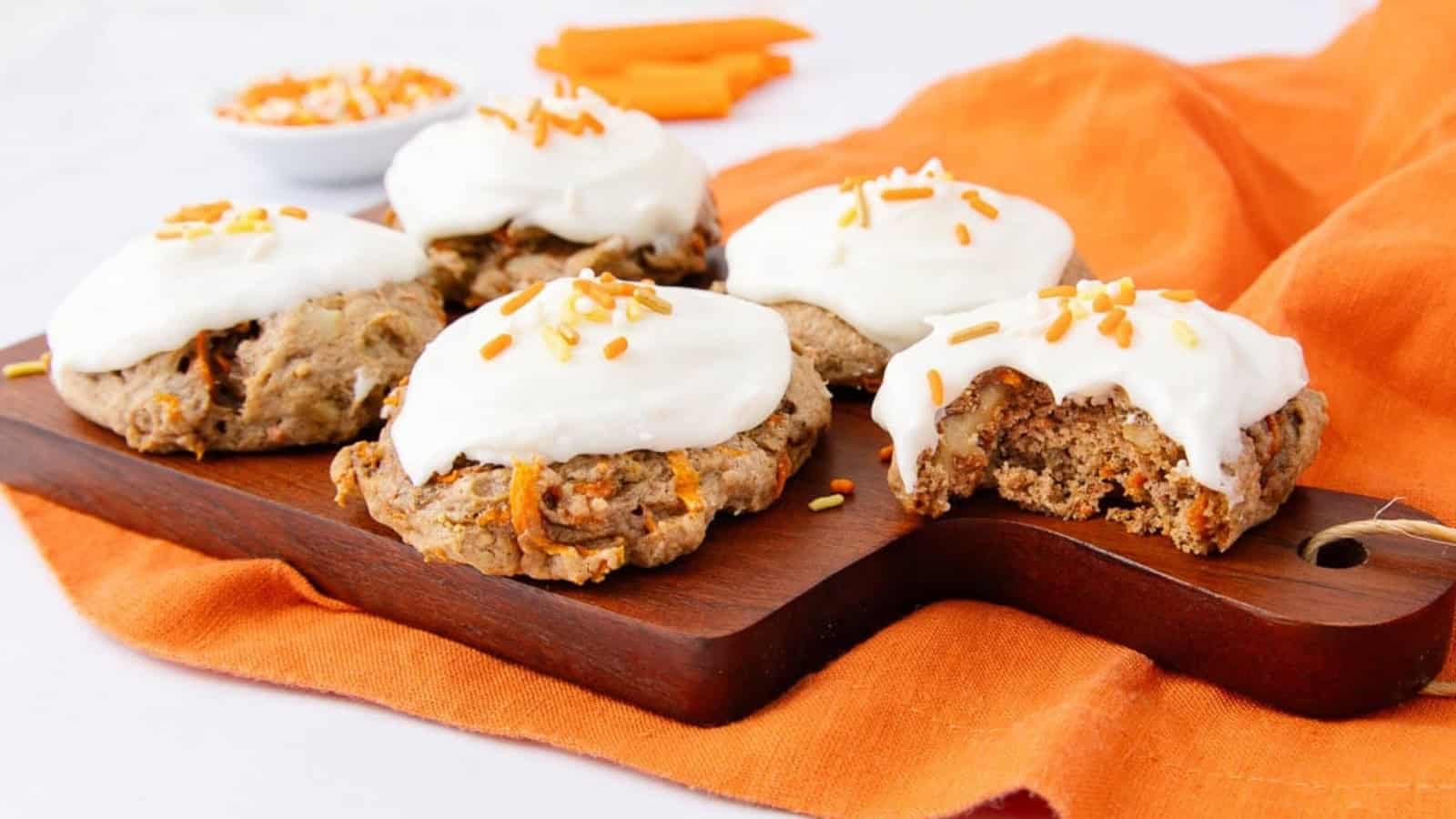 Carrot cake cookies with white icing and sprinkles on a wooden board, one with a bite taken out.