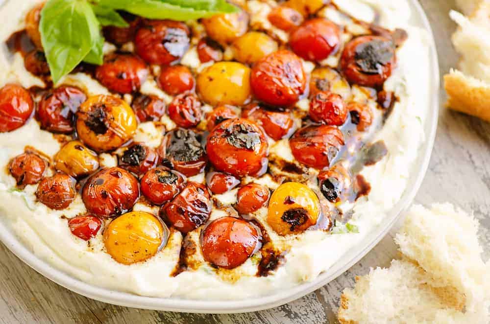 Roasted cherry tomatoes on creamy whipped cheese, drizzled with balsamic glaze, garnished with basil and bread.