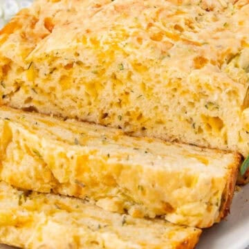 A loaf of cheesy bread with herbs, partially sliced, on a white plate with sprigs of rosemary.