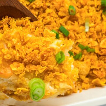A wooden spoon serving cheesy casserole topped with crispy cornflakes and sliced green onions—a classic among church potluck recipes.