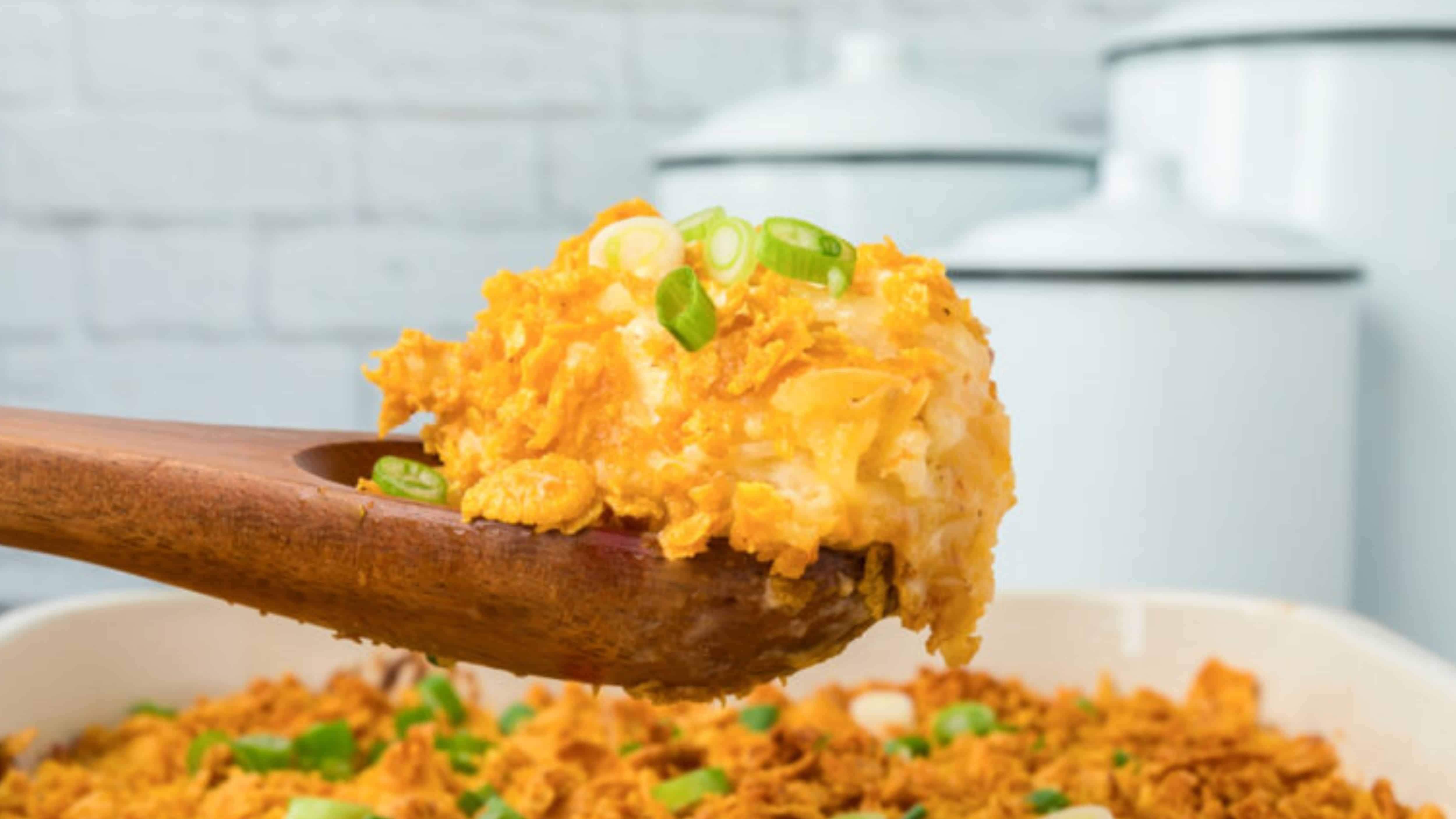 A wooden spoon holds cheesy, crunchy potato casserole topped with green onions above a baking dish.