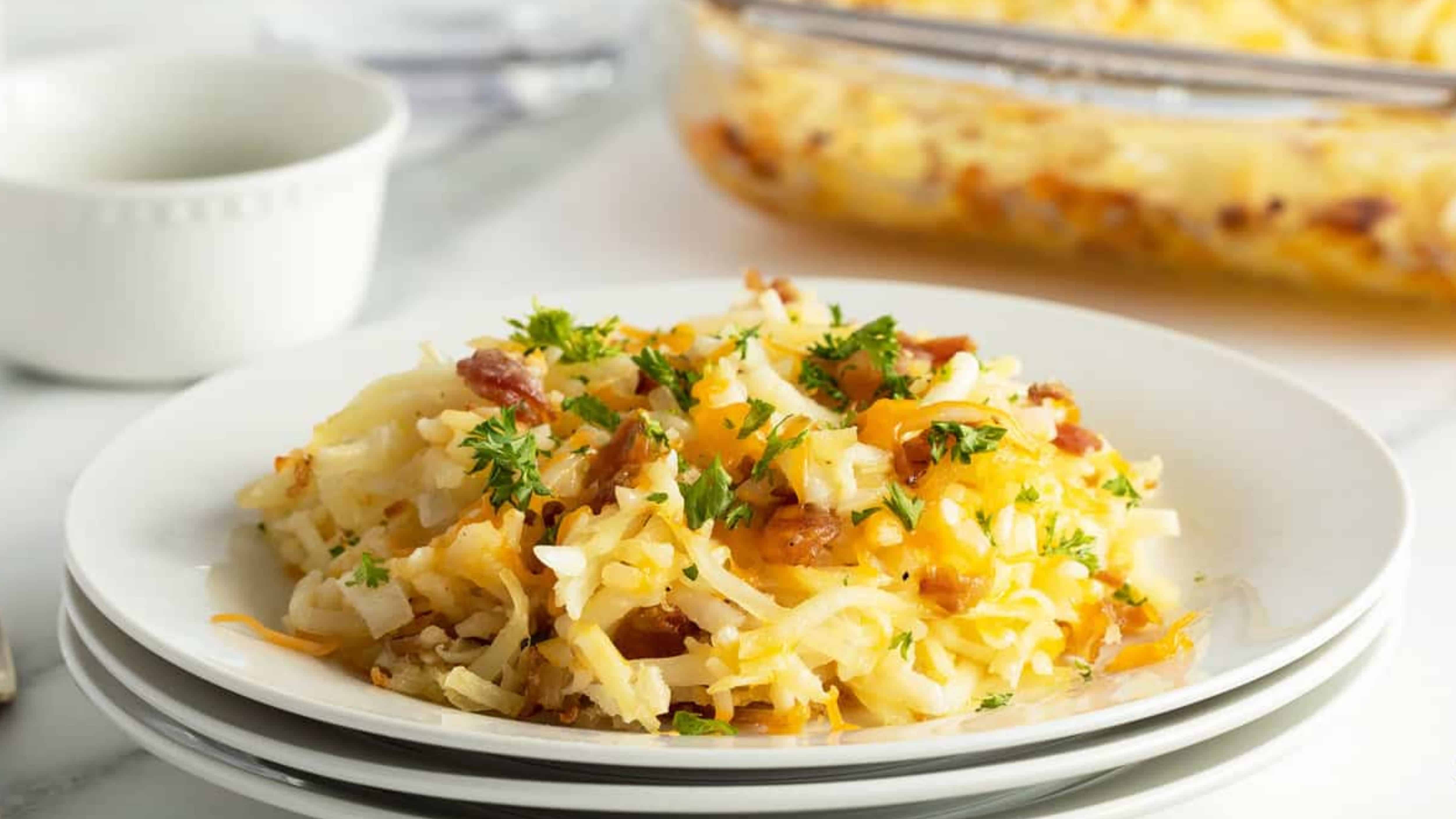 A serving of cheesy hash brown casserole with herbs on a white plate, casserole dish in the background.